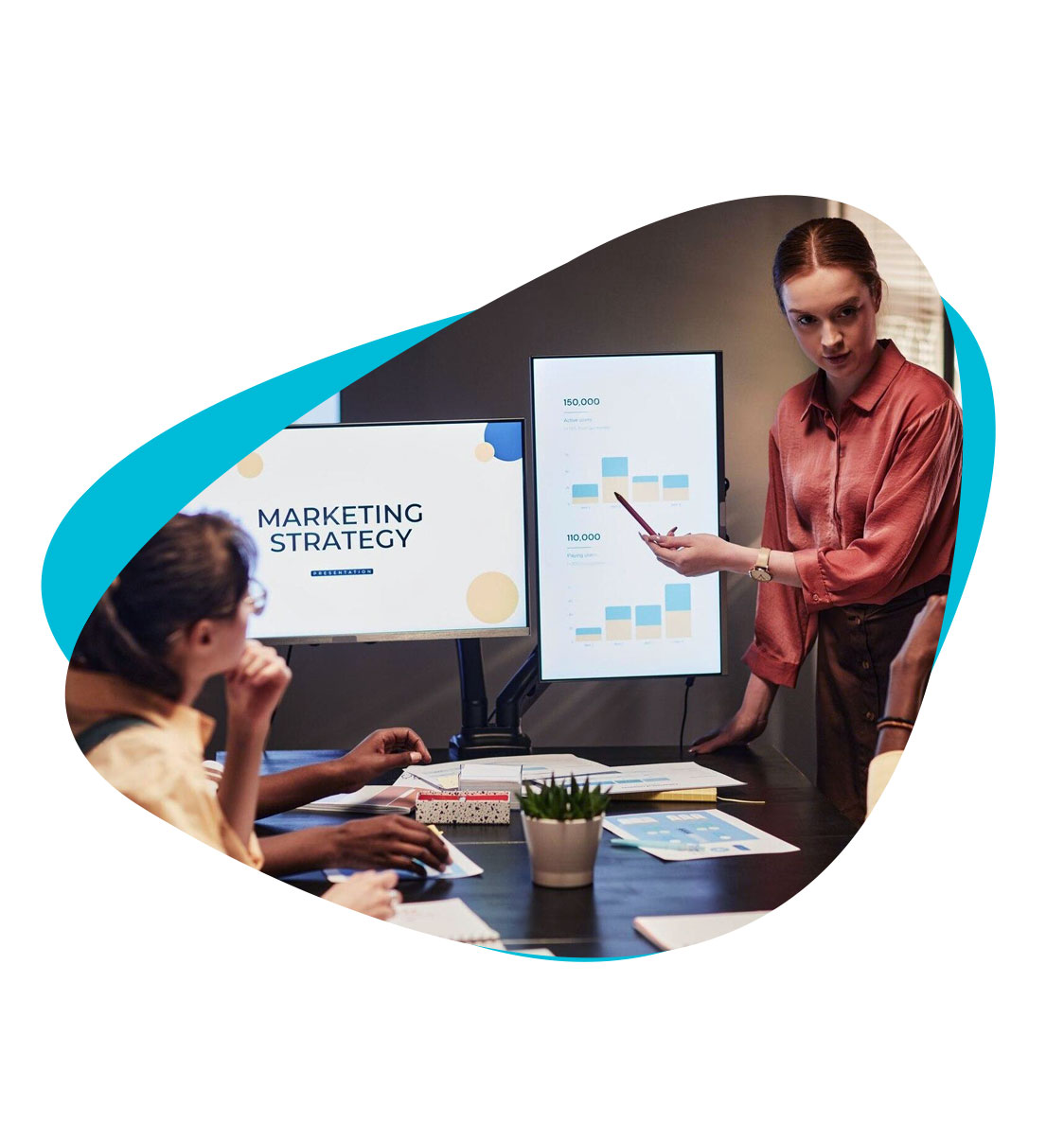 A woman presenting to a room of people. One of the monitors reads "Marketing Strategy." The other has graphs and charts.