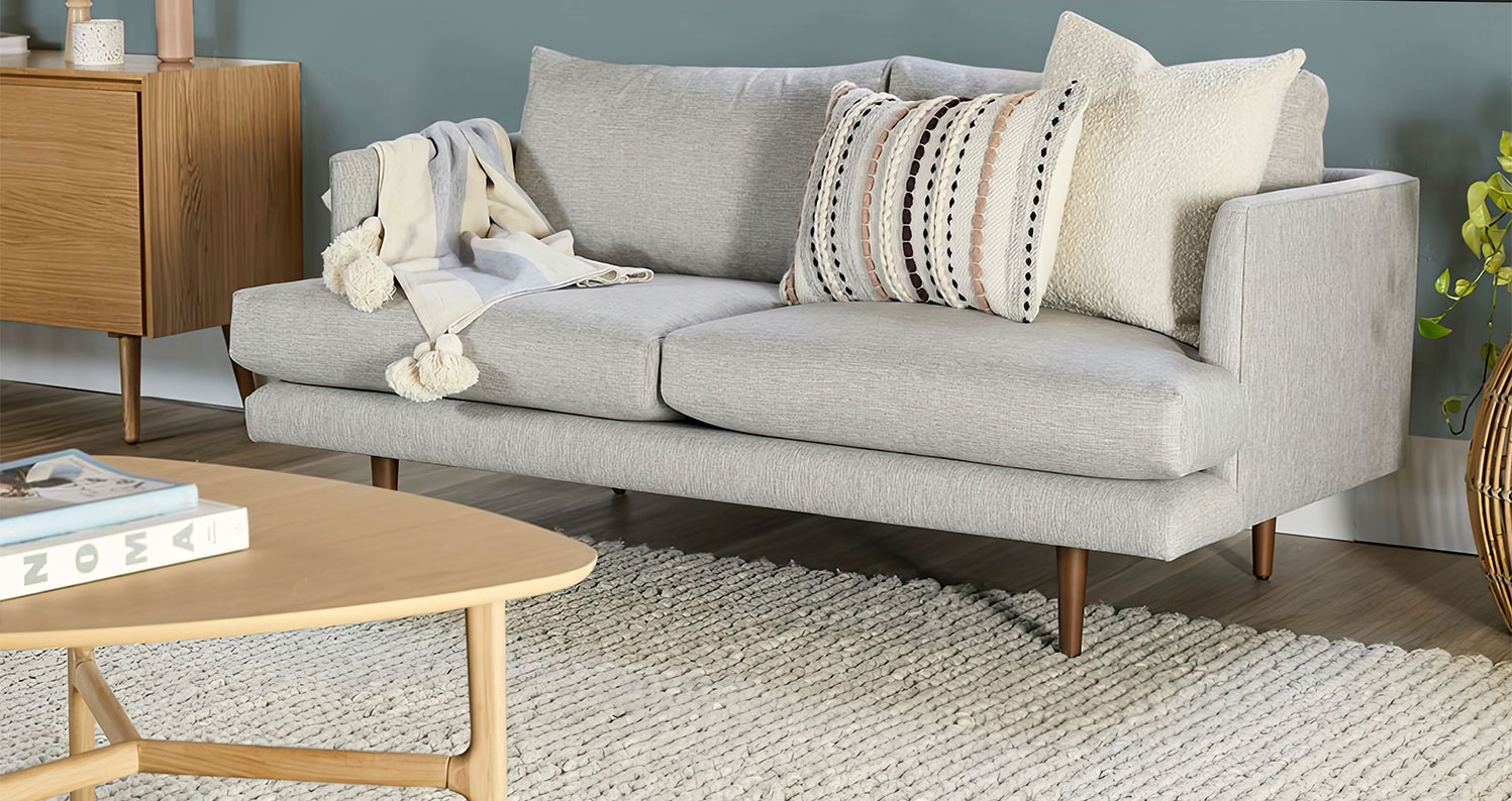 A modern, light gray sofa with mid-century wooden legs is adorned with textured cream and patterned throw pillows, and a tasseled blanket draped over one arm. A soft woven rug and a light wood coffee table with books add to the cozy, minimalist ambiance of the living room.