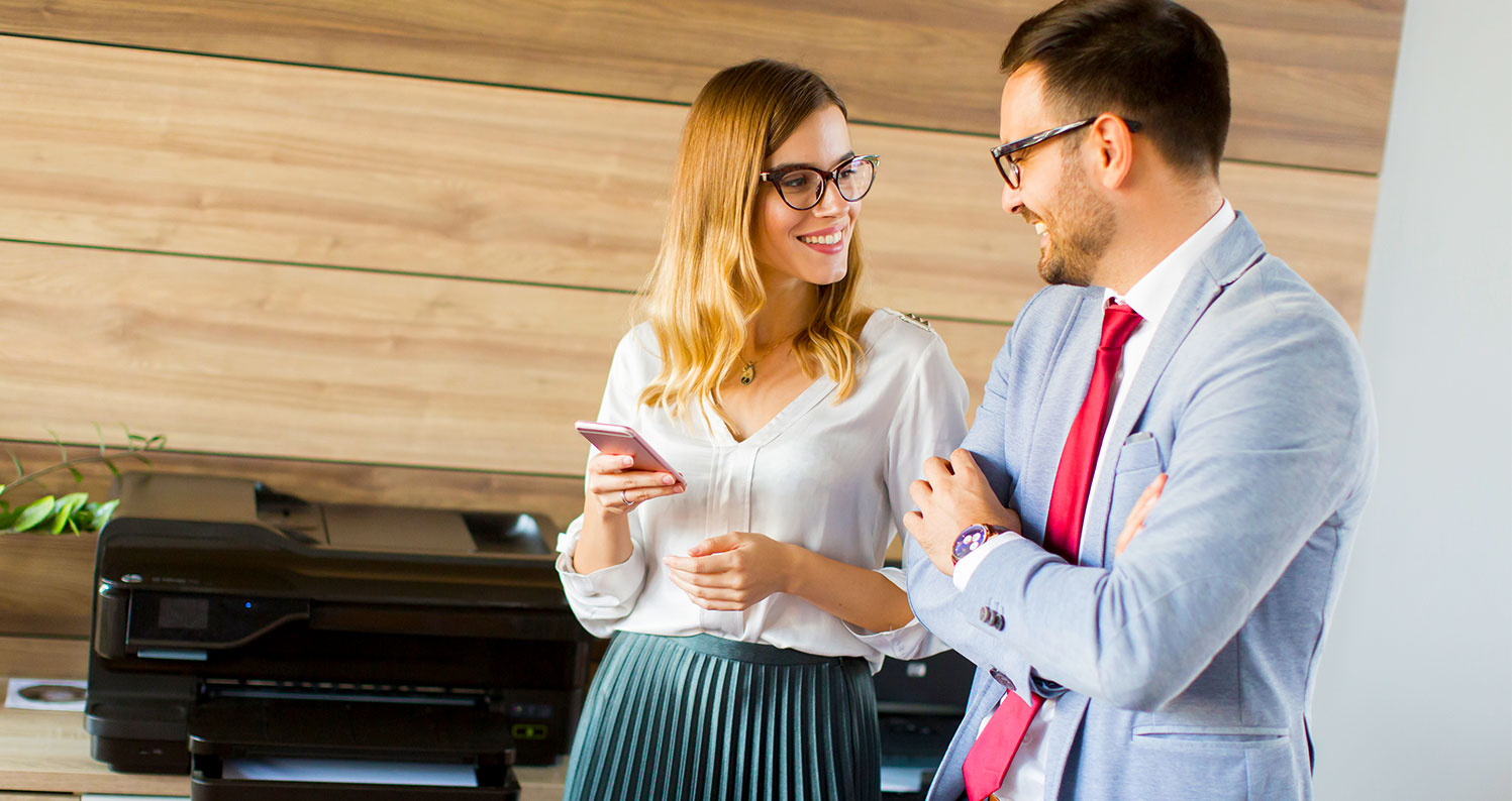 A woman holding a smartphone smiles while talking with a man in a light blue suit and red tie. They are standing in front of a printer in a modern office setting with wood panel walls.
